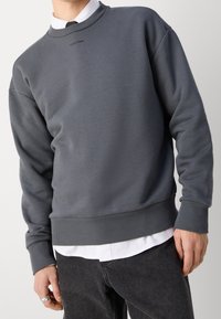 Felpa grigia a girocollo con polsini e orlo a coste, caratterizzata da un logo discreto sul petto. Indossata sopra una camicia bianca con colletto.