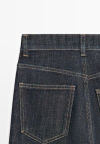 Donkerblauwe denimspijkerbroek met zichtbare achterste tailleband, riemlussen en twee achterzakken met oranje stikseldetails.