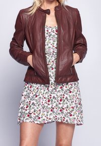 Mujer vestida con una chaqueta de cuero granate sobre un vestido floral blanco, con las manos en los bolsillos de la chaqueta, de pie contra un fondo liso.