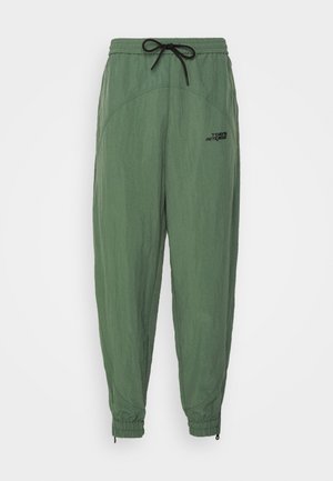 Veste légère verte et pantalon assorti, associés à une chemise orange à boutons et des baskets noires. Un sac fourre-tout jaune est visible.