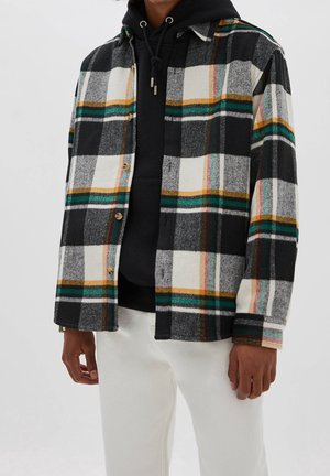 Męski model ubrany w koszulę na guziki w czarno-białą, zieloną i żółtą kratę, na czarnej bluzie z kapturem, oraz białe spodnie.