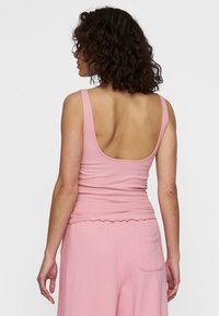 Vrouw met krullend donker haar draagt een mouwloze ribben top in het roze en losse roze broeken, met haar rug naar de camera gekeerd tegen een eenvoudige witte achtergrond.