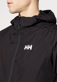 Schwarze Kapuzenjacke mit einem Frontreißverschluss, texturiertem Stoff und einem weißen Logo auf der linken Brust. Nahaufnahme des Kragens und des Reißverschlussdetails.