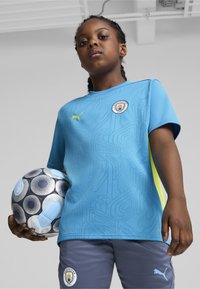 Vaaleansininen jalkapalloselkä, jossa on teksturoitu kuosi, keltaiset korostukset ja Manchester Cityn logo. Harmaat shortsit logolla ja pelaaja, joka pitää kuvioitua palloa.