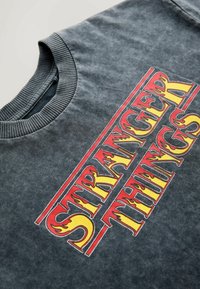 Tricou gri închis cu guler circular nervurat. Prezintă un grafic îndrăzneț "STRANGER THINGS" în flăcări roșii și galbene în centrul său.