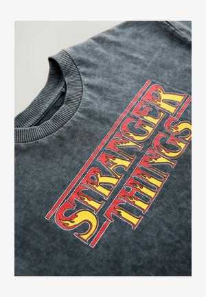 Tamno sivi majica s rebrastim okruglim izrezom. Sadrži hrabre grafike "STRANGER THINGS" u crvenim i žutim plamenima na sredini.