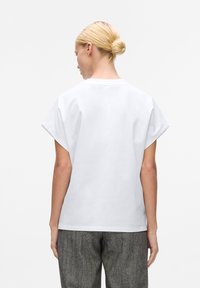 Tricou alb din bumbac, cu mâneci scurte tip dolman, decolteu rotund și textură netedă; purtat cu pantaloni gri cu model. Vedere din spate.