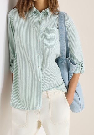 Femme portant une chemise à rayures vert clair boutonnée et un pantalon blanc cassé, portant un sac en tissu bleu clair sur l'épaule.