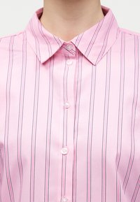 Roosa triiksärk vertikaalsete mustade ja valgete triipudega, millel on terav krae ja esiküljel nähtavad nupud.