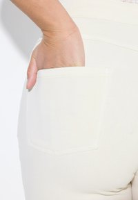 Bílé denimové džíny s viditelnou zadní kapsou, s jemnou texturou a detaily šití. Ruka spočívá uvnitř kapsy.