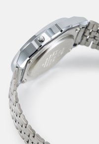 Montre en métal argenté avec un bracelet en acier inoxydable, boîte octogonale, couronne à bouton-poussoir et finition lisse brossée au dos.