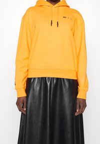Sudadera corta naranja con cordones y logo negro, combinada con una larga falda de cuero negra. Textura lisa, diseño informal.