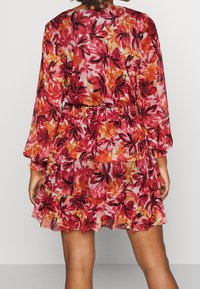 Vestido floral en rojo y naranja con cintura fruncida, mangas largas, volantes en capas y tela translúcida. Presenta un patrón vibrante y multicolor.