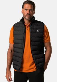 Schwarze, gefütterte Weste mit hohem Kragen, gestepptem Muster und durchgehendem Reißverschluss, getragen über einem orangen Polohemd. Mit Logo auf der linken Brust.