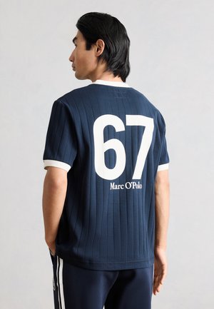 Uomo visto di profilo che indossa una maglietta a coste blu navy con la scritta bianca "67" e "Marc O'Polo" sulla schiena, in piedi davanti a uno sfondo semplice.