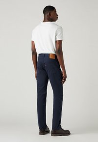 Marinblå byxor med rak bendesign, med en brun läderlapp på midjan och dubbla bakfickor. Vit t-shirt bärs.