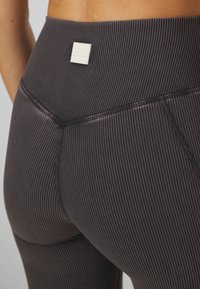 Nära bild av en person i mörkgrå högmidjade leggings med tunna vertikala vita ränder och en liten vit PE Nation-logotyp på midjebandet.