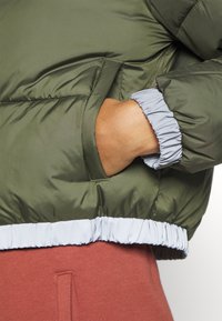 Giacca puffer verde oliva con polsini elastici e orlo grigio chiaro. Presenta una tasca laterale e una texture esterna liscia e lucida.
