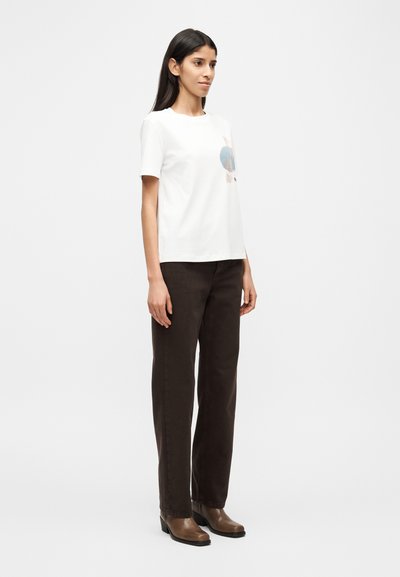 Λευκό κοντομάνικο μπλουζάκι με μπλε και καφέ γραφικό σχέδιο, συνδυασμένο με σκούρες καφέ παντελόνες ίσιας γραμμής και καφέ μποτάκια με τακούνι.