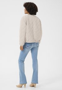 Veste bomber beige matelassée avec un col arrondi, un ourlet élastiqué et des manches longues, associée à un jean évasé bleu clair et des talons beiges.