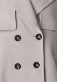 Cappotto doppiopetto realizzato in morbido tessuto grigio chiaro. Presenta grandi bottoni neri e colli dettagliati, con una finitura liscia e strutturata.