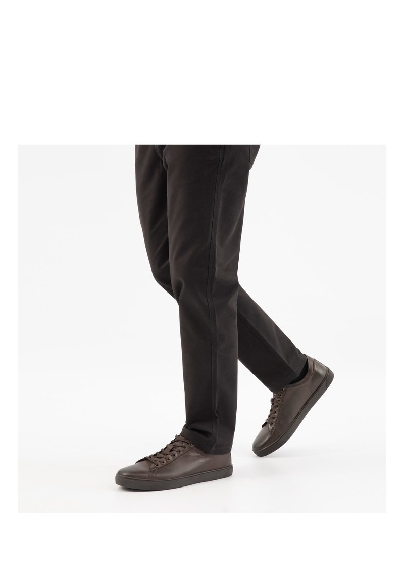 Man draagt zwarte broek en donkerbruine leren sneakers, lopend op een effen witte achtergrond.