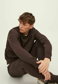 Bruine sweater en bijpassende joggingsbroek van zachte stof. De outfit heeft ribgebreide boorden en een relaxte pasvorm. De achtergrond is licht en neutraal.