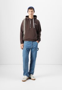 Brown hooded sweatshirt with white drawstrings, blue jeans with curved pockets, and white sneakers with brown soles. Grey shoulder bag.