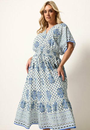 Donna che indossa un vestito bianco con motivi floreali e geometrici blu, maniche corte, scollo a V e vita elastica, in piedi davanti a uno sfondo uniforme.