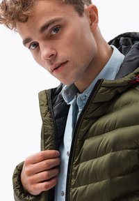 Giacca imbottita verde oliva con fodera nera, dotata di chiusura con zip e tasche laterali, indossata sopra una camicia azzurro chiaro con colletto.