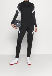 En person som har på sig svart och vit Nike sportkläder håller en svart och grå fotboll och står framför en helt vit bakgrund.