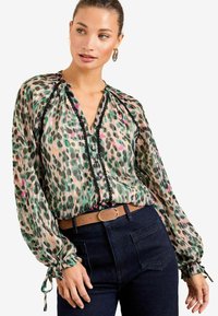 Caurs, rakstainais blūze ar zaļiem, rozā un melniem punktiem, gariem piedurknēm ar elastīgām aprocēm un pogājamo priekšpusi ar melniem akcentiem.