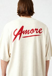 Kremi puuvillane oversized T-särk, mille seljal on suur punane kaldkiri "Amore". Lühikesed varrukad ja mugav lõige.