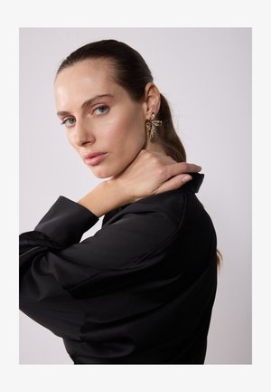 Schwarze Satinbluse mit langen Ärmeln und strukturiertem Kragen. Goldene, blumige Ohrringe setzen Akzente. Heller Hintergrund, neutrale Farbtöne.