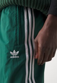 Zelené sportovní kalhoty s bílými bočními pruhy, obsahující zipovou kapsu a vyšitým logem Adidas na levém stehně. Materiál vypadá lehce.