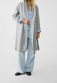 Persona vestida con un abrigo largo gris, camisa a rayas azul, jeans anchos claros y rasgados, zapatos negros y anillos de oro en los dedos.