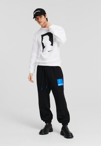 Weißer Sweatshirt mit einem schwarzen Silhouettenprint, kombiniert mit schwarzen Sweatpants, die ein blaues Logopatch und einen Kordelzug haben, sowie schwarzen Stiefeln.