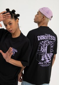Twee mensen dragen zwarte "Discoteca" shirts; de één staat frontaal met opgeheven handen, de ander toont de achterkant van het shirt met een discothema en een paarse pet.
