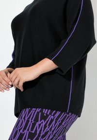 Czarna dzianinowa bluzka z fioletowym wykończeniem na rękawach, zestawiona z fioletowymi legginsami z wyraźnymi czarnymi literami. Gładka struktura, luźny krój.