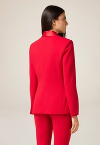 Czerwona, dopasowana marynarka o wąskim kroju z satynowym kołnierzem. Gładka tkanina i czyste linie, uzupełnione pasującymi spodniami.
