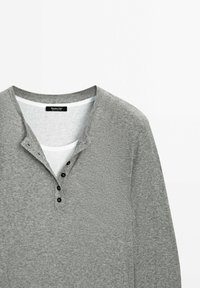 Maglietta grigia a maniche lunghe tipo henley con tessuto testurizzato, tre bottoni neri sul collo e una camicia bianca visibile sotto il colletto.