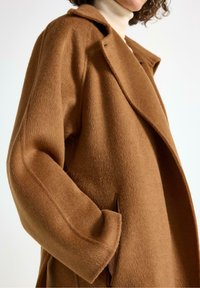 Cappotto di lana marrone con colletto a punta, maniche ampie e tasche laterali. La texture è morbida, offrendo un'aspetto caldo e accogliente.