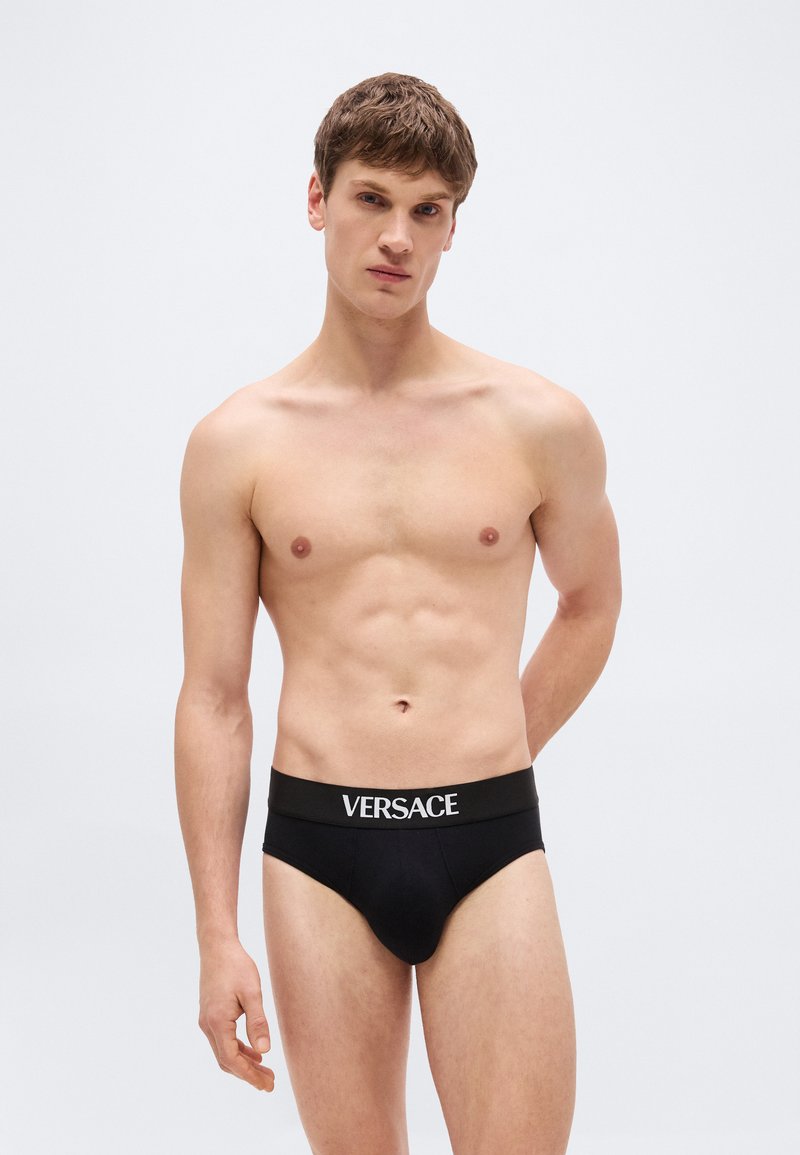 Young man standing, wearing black Versace briefs, neutral expression, against plain light gray background.