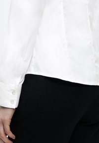 Λευκό πουκάμισο με μακριά μανίκια που διαθέτει απαλή υφή, μανσέτες με κουμπιά και εφαρμοστή ραφή, συνοδευόμενο από μαύρο παντελόνι από ύφασμα.