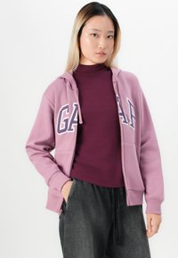 Lichtroze zip-up hoodie met het "GAP" logo, zachte textuur, gedragen over een aansluitend bordeaux top en gecombineerd met losse grijze broek.