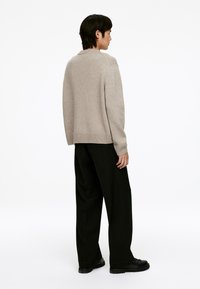 Beiger Strickpullover mit geripptem Saum und Bündchen, kombiniert mit hochtaillierten schwarzen Hosen und schwarzen Schuhen, von hinten betrachtet.