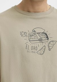 Beige cotton t-shirt featuring a black graphic illustration of a sardine tin and citrus slices, with the text "EL ORO DEL MAR" below.