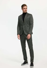 Traje gris texturizado con un diseño ajustado, combinado con un cuello de tortuga negro y zapatillas negras. Presenta solapas con muesca y pantalones de frente plano.