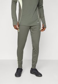 Tamnozeleni sportski outfit s dugim rukavima i prilagođenim hlačama s svijetlozelenim akcentima. Materijal izgleda rastezljivo i glatko.