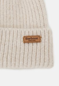 Căciulă tricotată bej, cu design cu nervuri, având o etichetă din piele maro pe care scrie "Barbour ENGLAND." Textură moale și grosime medie.